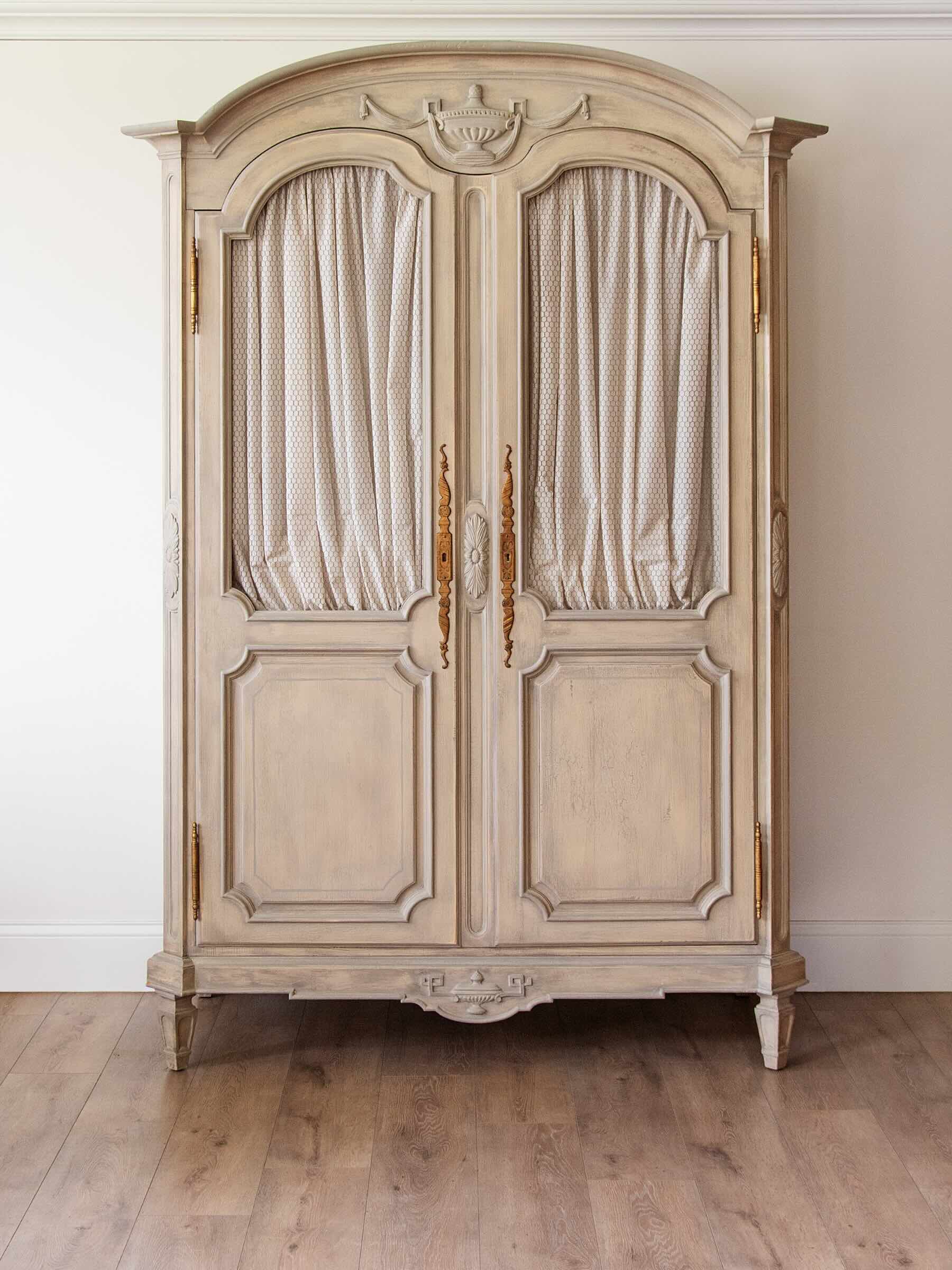 A large vintage-style painted wooden armoire cabinet with two arched door panels, draped cream curtains, ornate gold handles, and a distressed off-white finish, set against a plain wall and light wood floor.