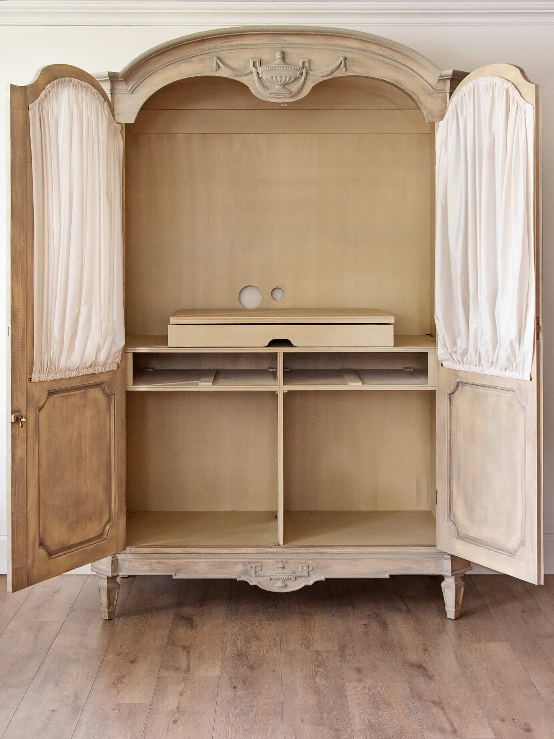 A large, vintage-style wooden armoire cabinet with its doors open, showing cream-colored interior curtains, shelves, and a top drawer. The cabinet has a light beige and brown painted finish, set against a neutral wall and a wooden floor.