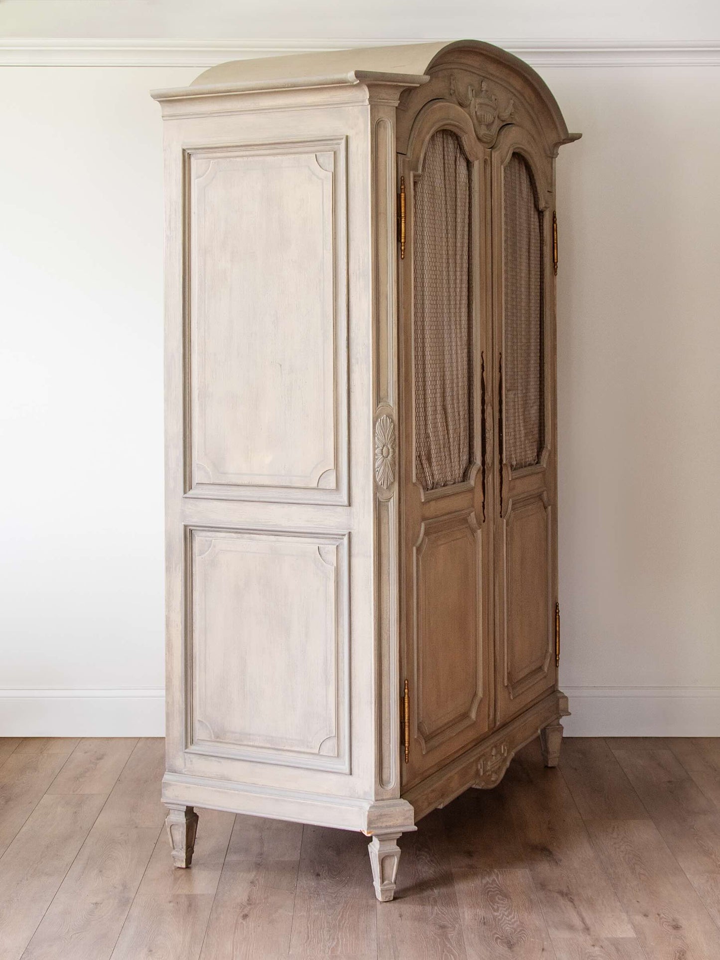 A tall vintage-style wooden armoire cabinet with a distressed finish, featuring one panel door and one double door with woven cane inserts, set against a plain white wall and light wood floor.