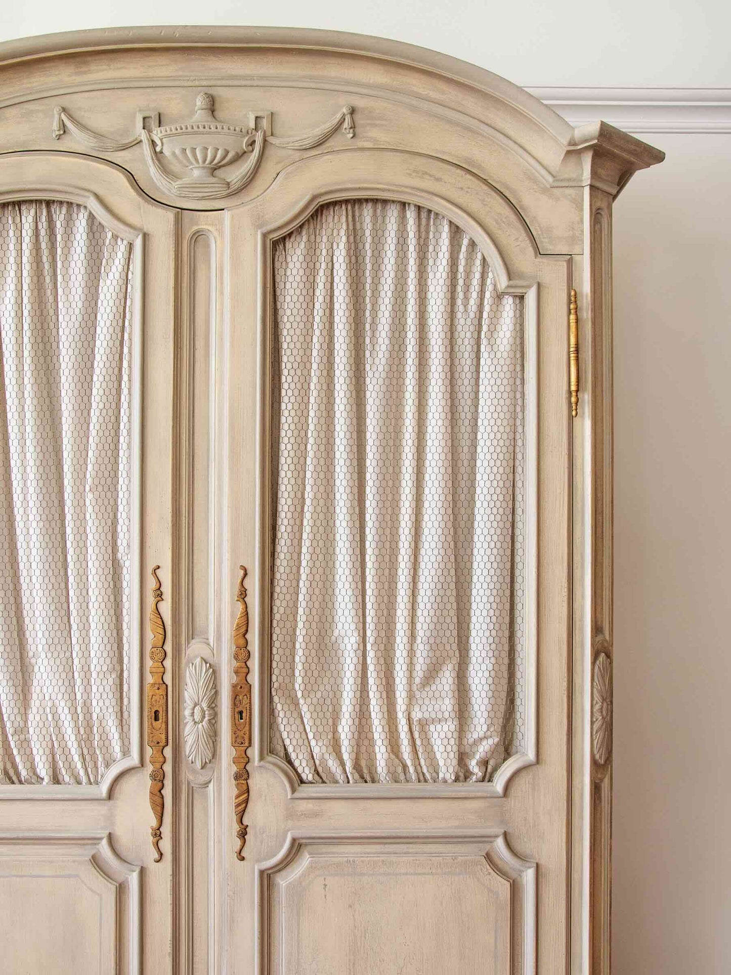 An ornate vintage-style armoire cabinet in a light beige/cream finish with gold-toned handles and brass hinges, featuring patterned cream curtains in the door panels.