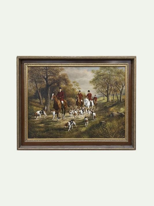 A framed landscape painting depicting a fox hunting scene with riders on horseback and hounds in a wooded clearing, shown against a plain light background.