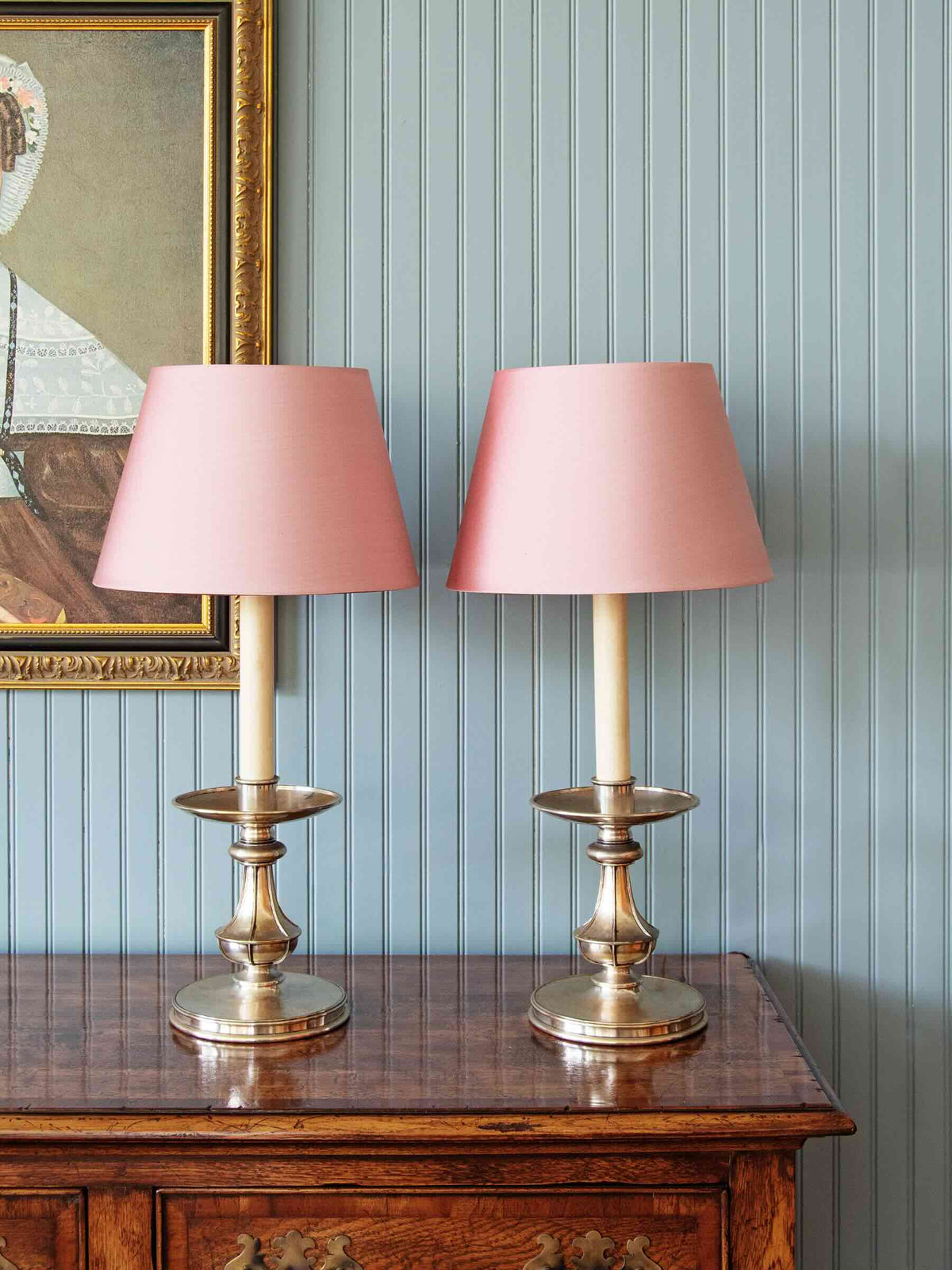 Two matching table lamps with dusty pink drum shades and antique brass bases, displayed side-by-side on a polished wooden console table against a light blue paneled wall.