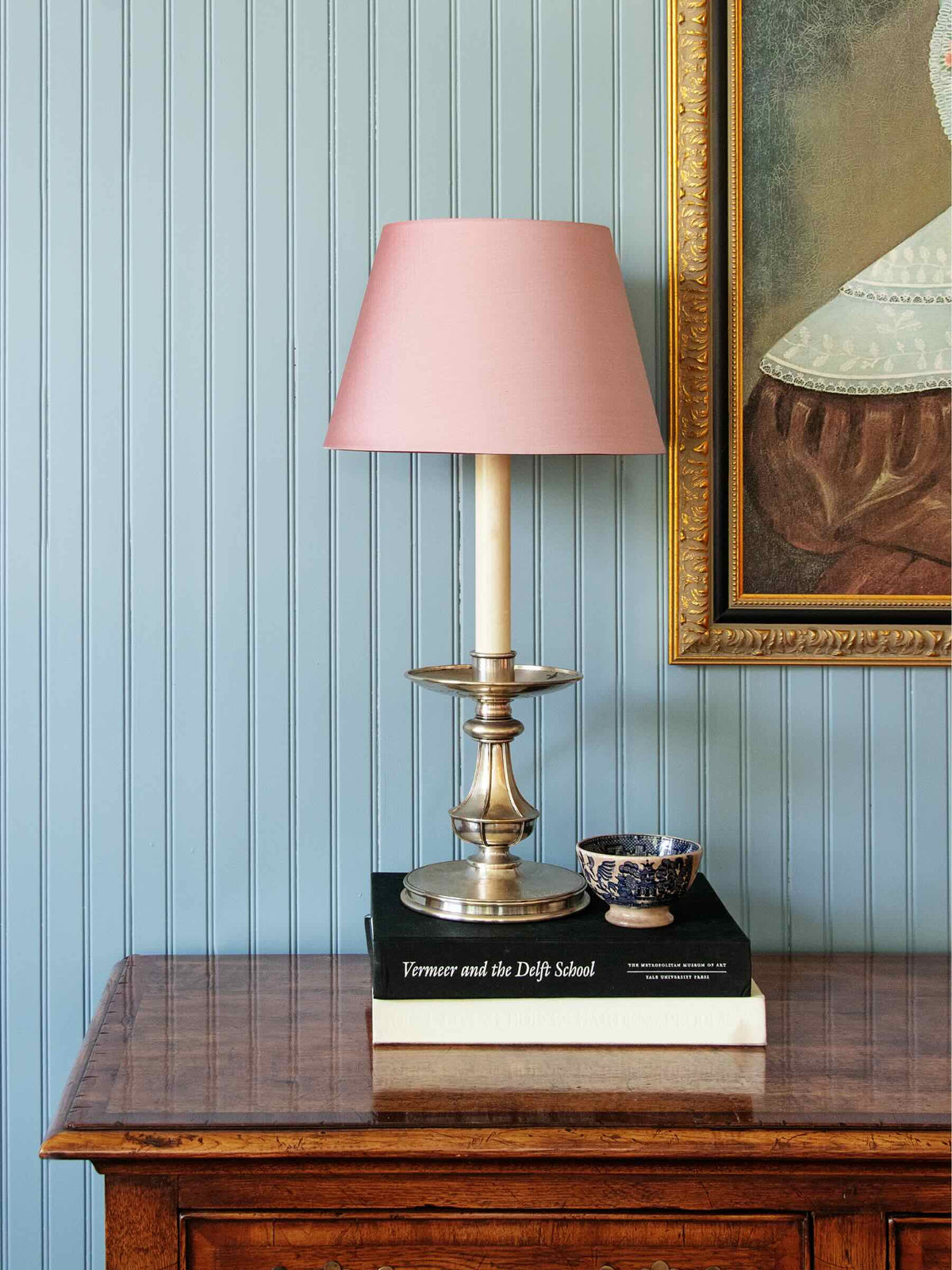 A table lamp with a dusty pink shade and a metallic gold/brass base sits on stacked books on a wooden console table against a blue-beadled wall.