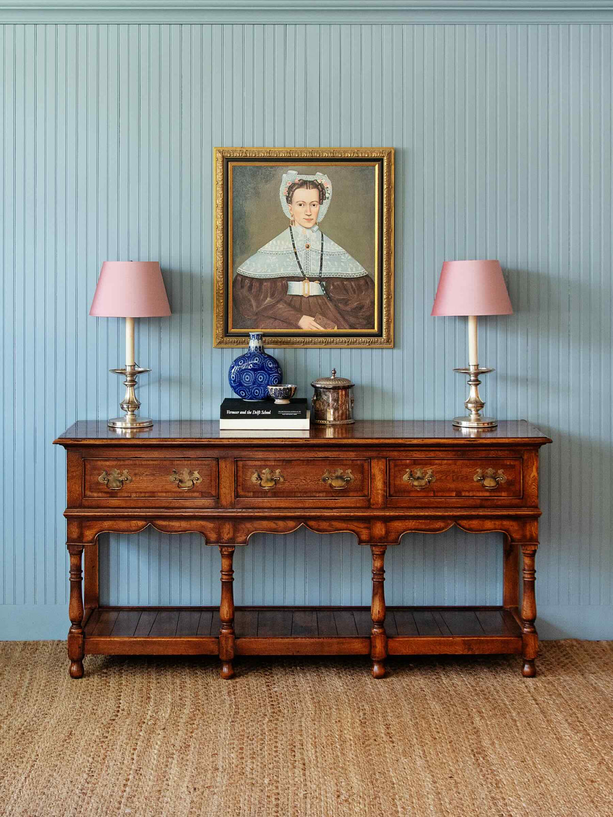 An ornate dark brown wooden console table with three drawers and a lower shelf, styled with two pink lampshades, a blue glass bottle, and a framed portrait on a light blue paneled wall.