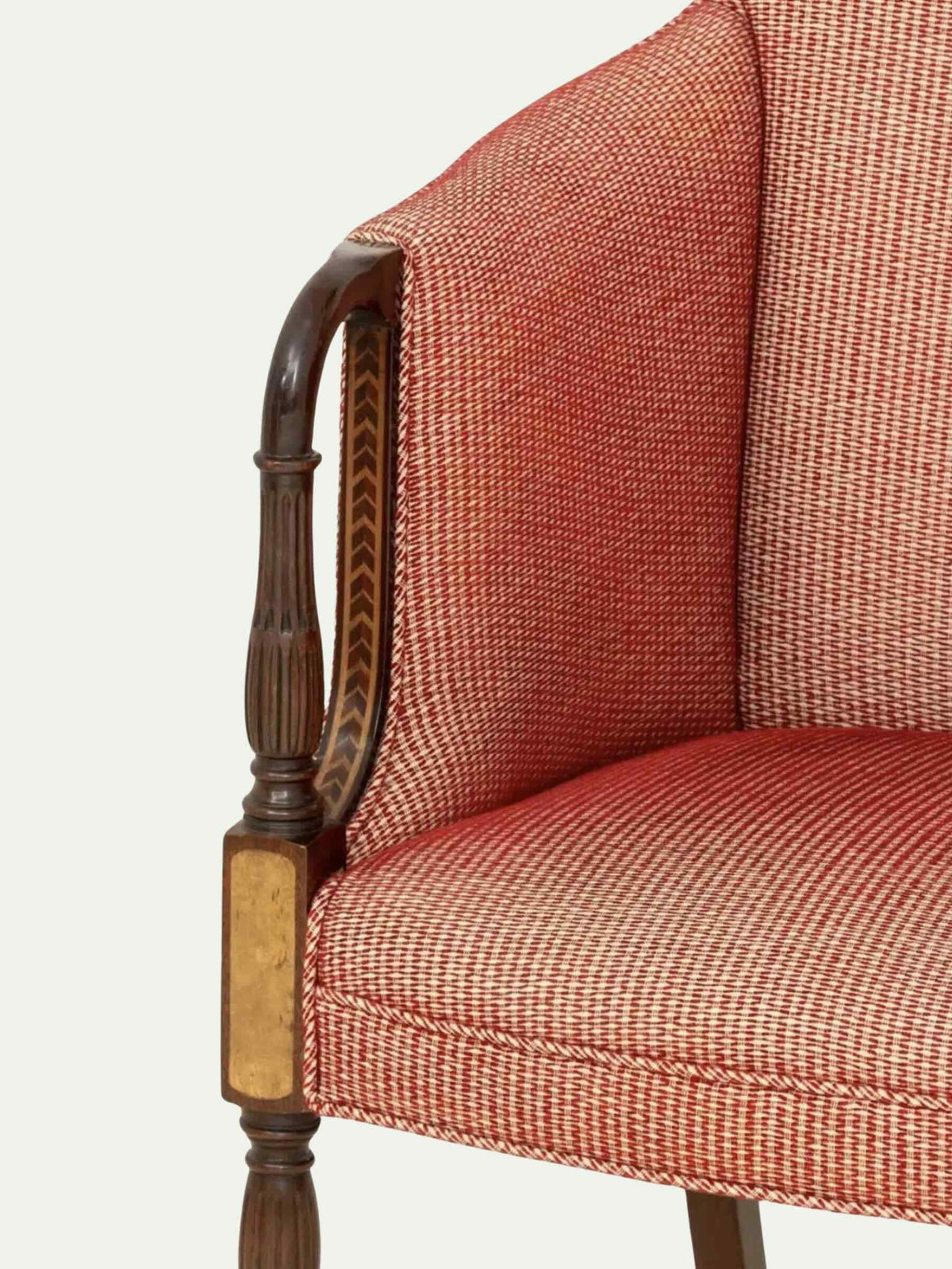 Close-up of an upholstered armchair corner showing textured rust-red fabric with a subtle woven pattern and dark wood armrest with brass-toned accents, set against a plain light background.