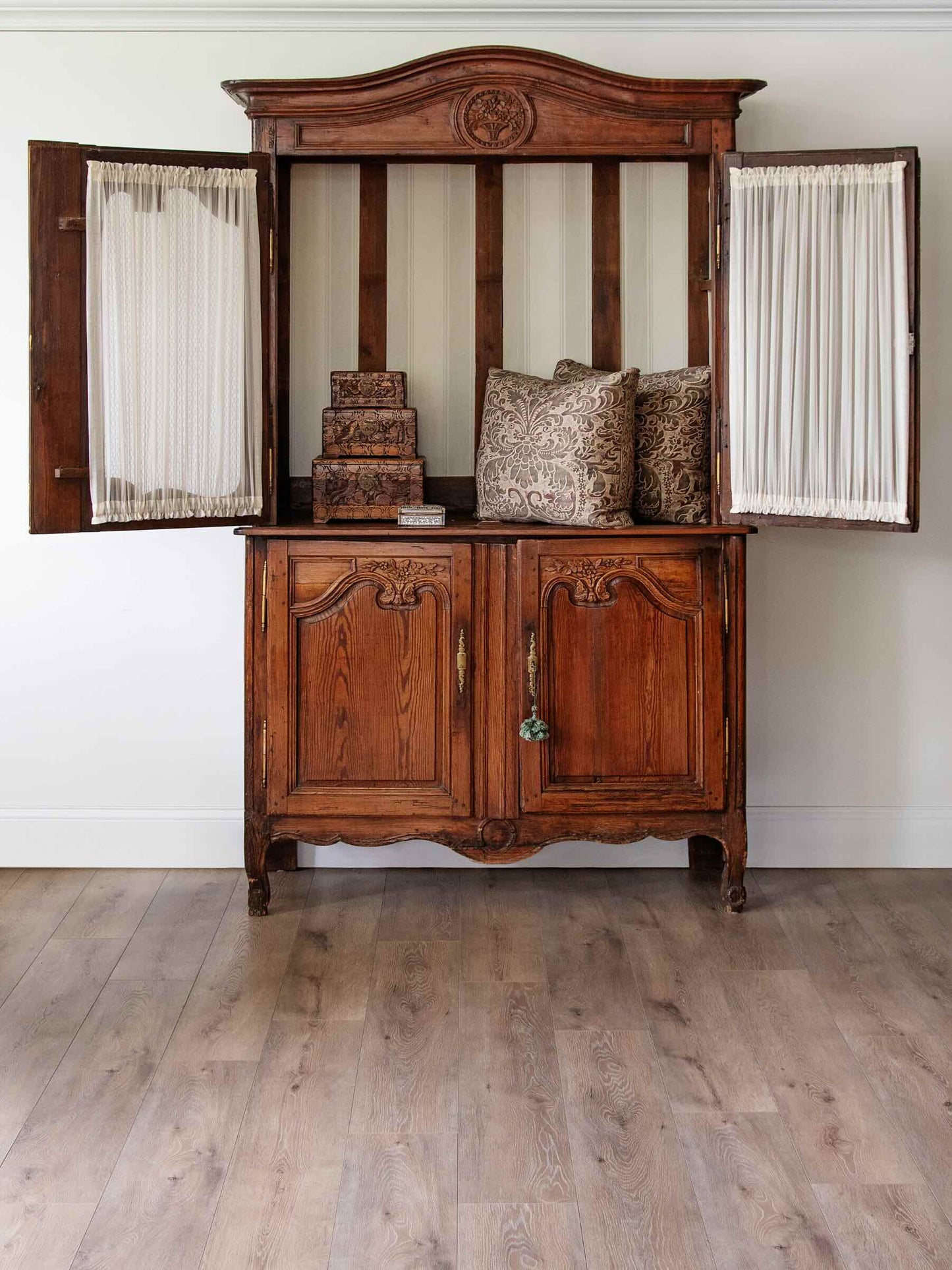 An antique dark brown wooden hutch or cabinet with open side doors, cream curtains, and decorative pillows on top, set against a white wall and light wood floor.