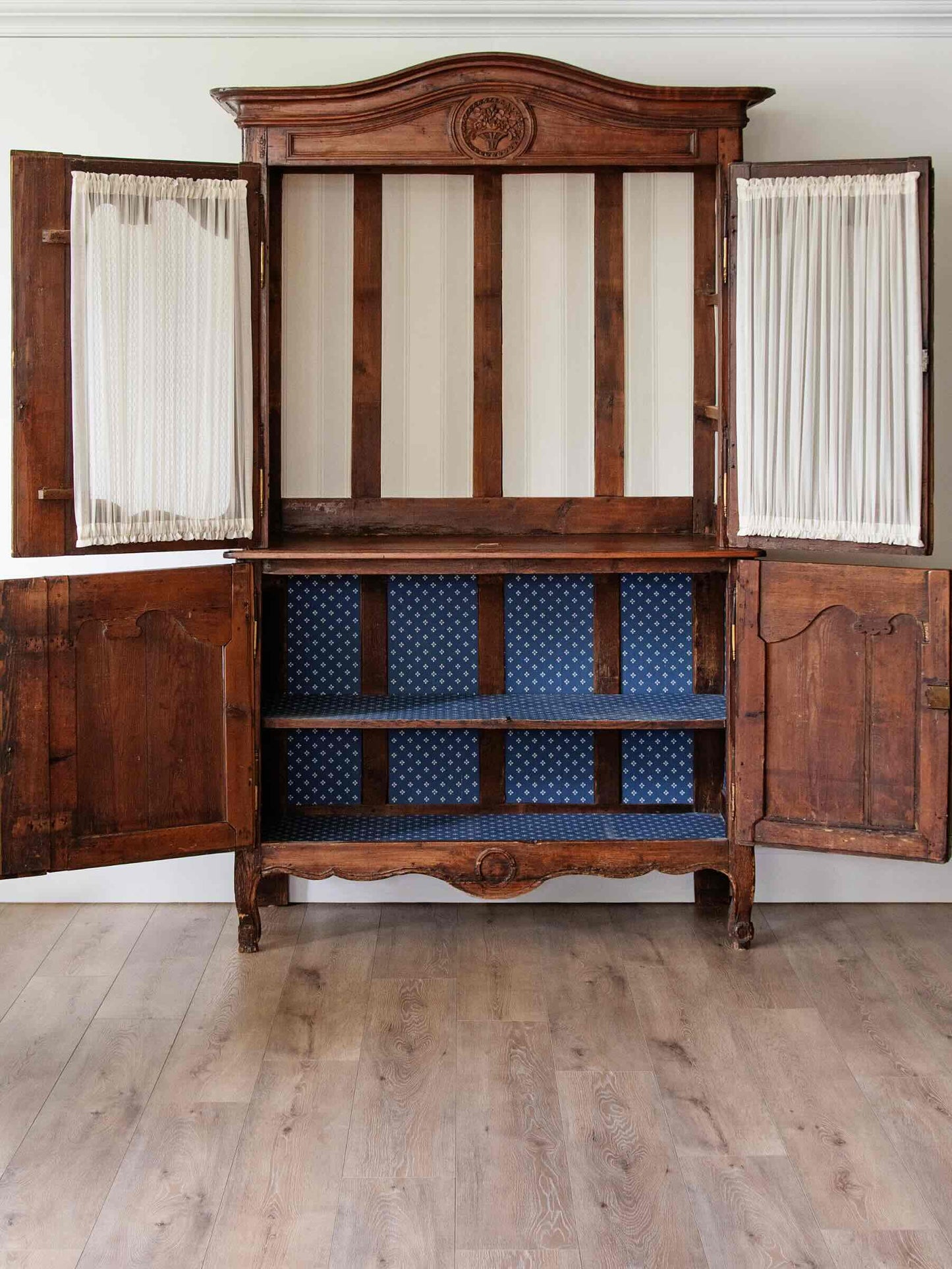 A large antique wooden hutch or cabinet with open doors, cream pleated curtains on the upper panels, and blue patterned fabric lining the lower shelves, set against a white wall and light wood floor.
