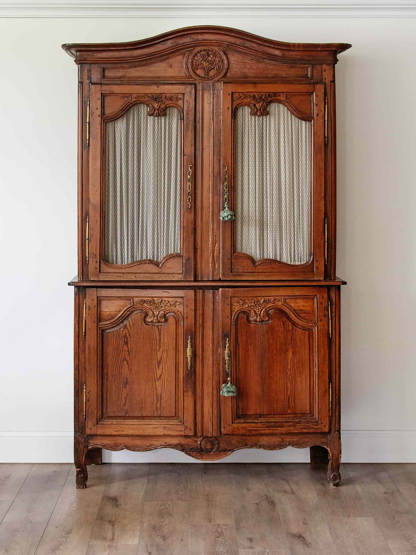 An antique French-style wooden hutch with carved details, featuring two upper doors with striped fabric inserts and two lower wooden doors, set against a plain white wall and light wood floor.