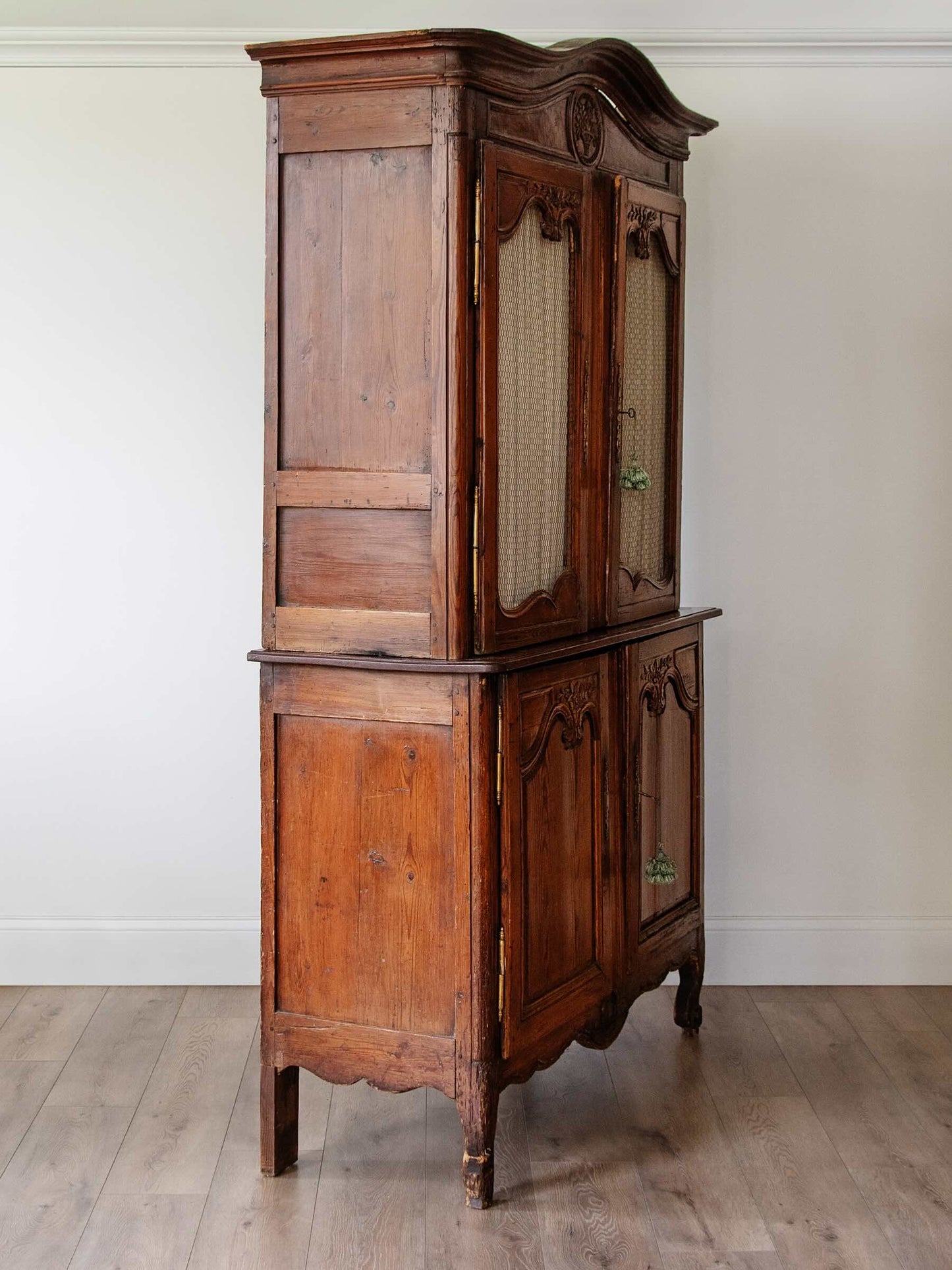 An antique wooden hutch or cabinet with a warm medium-brown finish, featuring two upper doors with woven cane panels and two lower cabinet doors, set against a plain white wall and light wood floor.