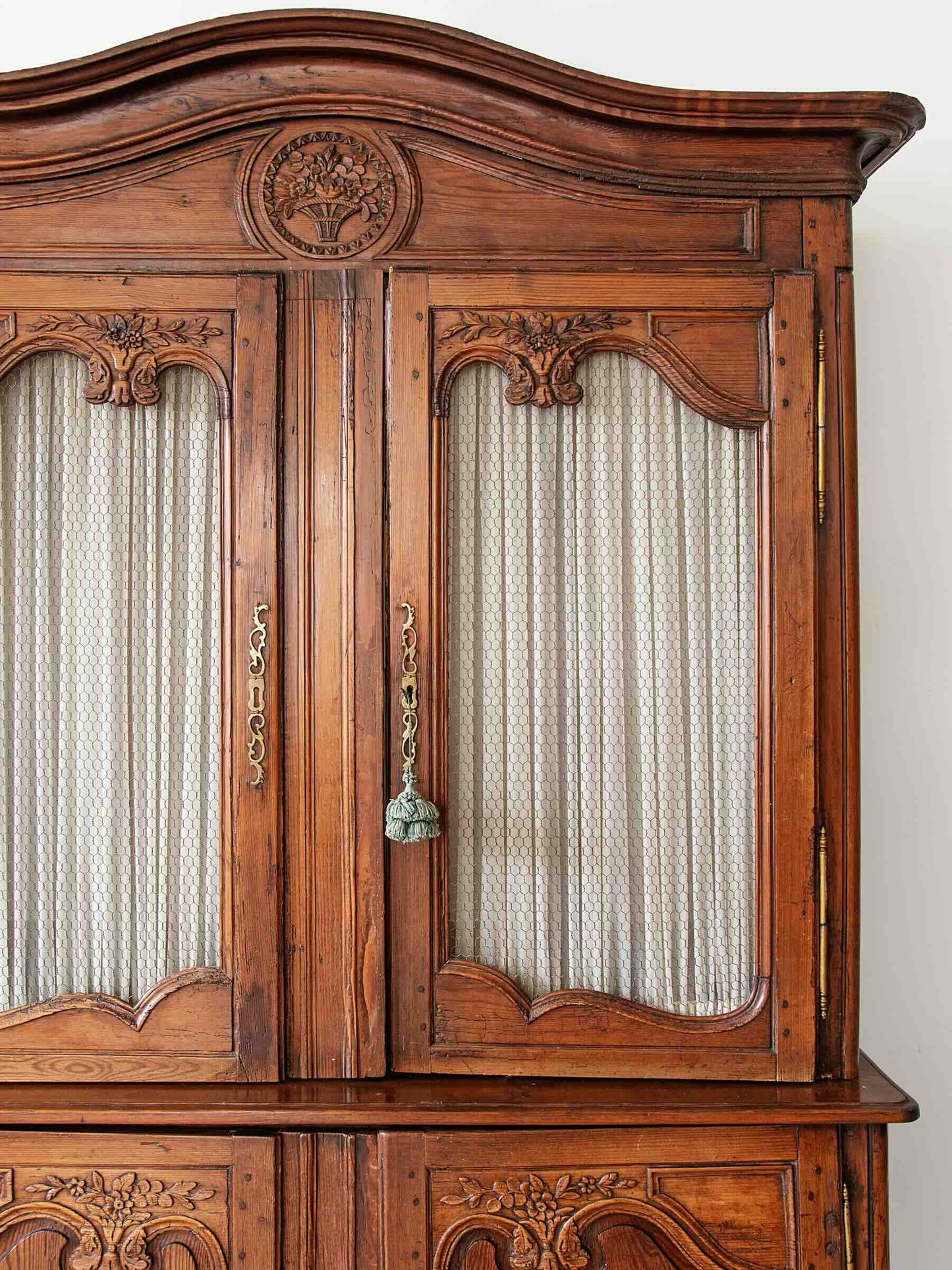 An ornate vintage wooden hutch or cabinet with carved details, featuring two glass-front doors with cream striped curtains and brass handles.