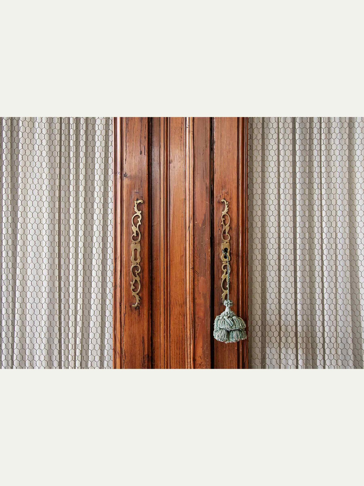 Close-up of a wooden door with ornate brass handles and a hanging tassel, flanked by patterned curtains on a light gray background.