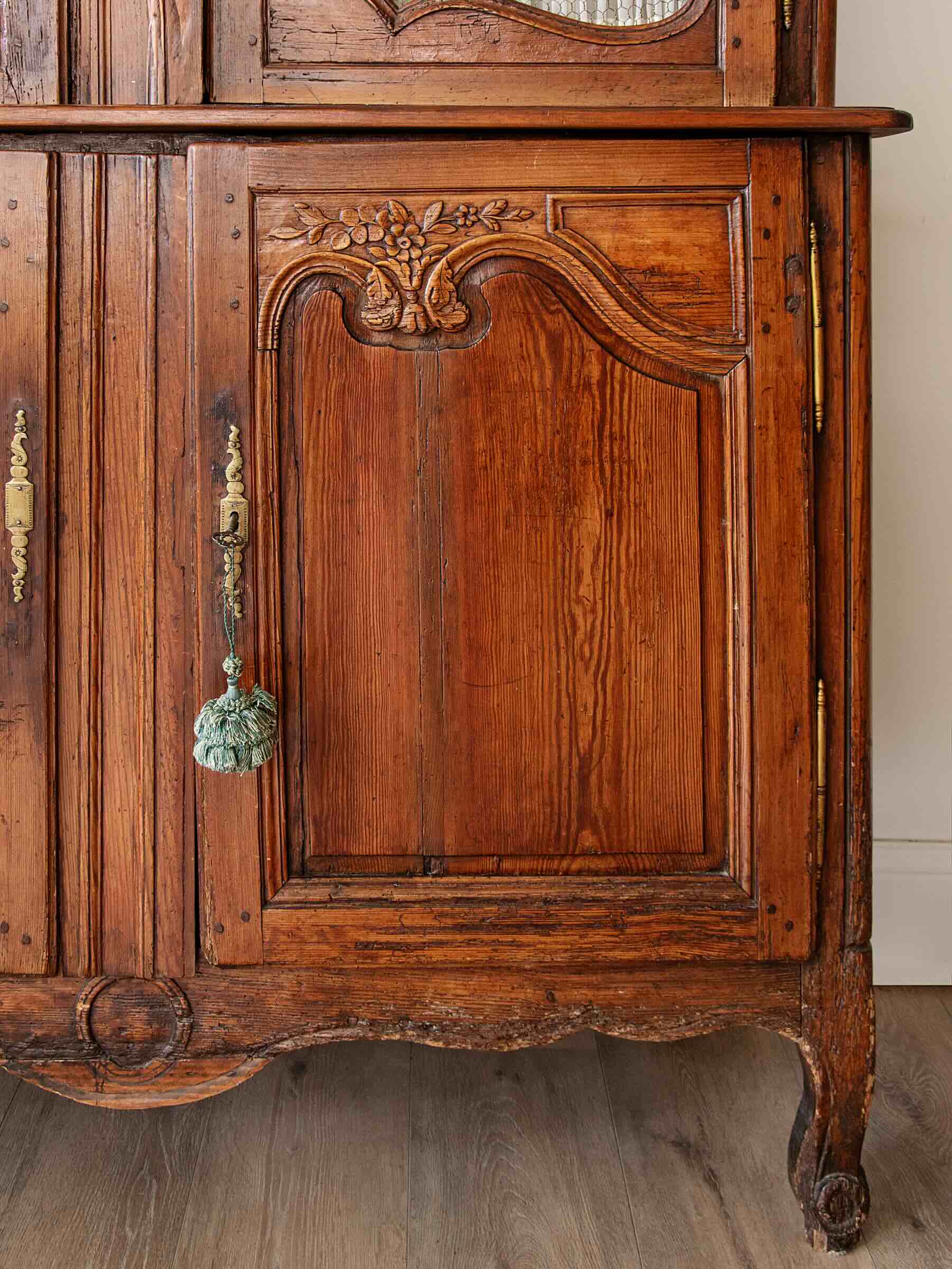 Close-up of an antique carved wooden cabinet with a warm brown finish, brass hardware, and a decorative hanging pull, shown against a light wall and wood floor.