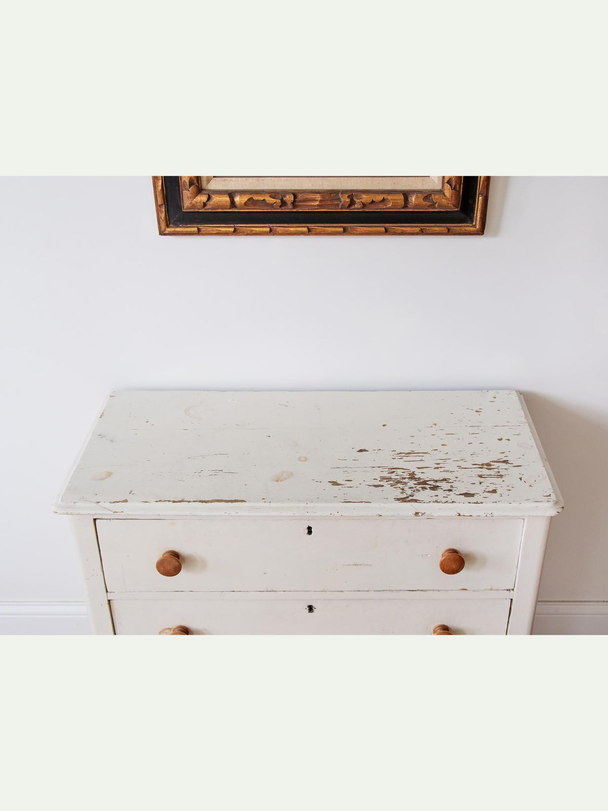 A distressed white wooden chest of drawers with two drawers and round wooden knobs, set against a white wall with a gold-framed mirror partially visible above.