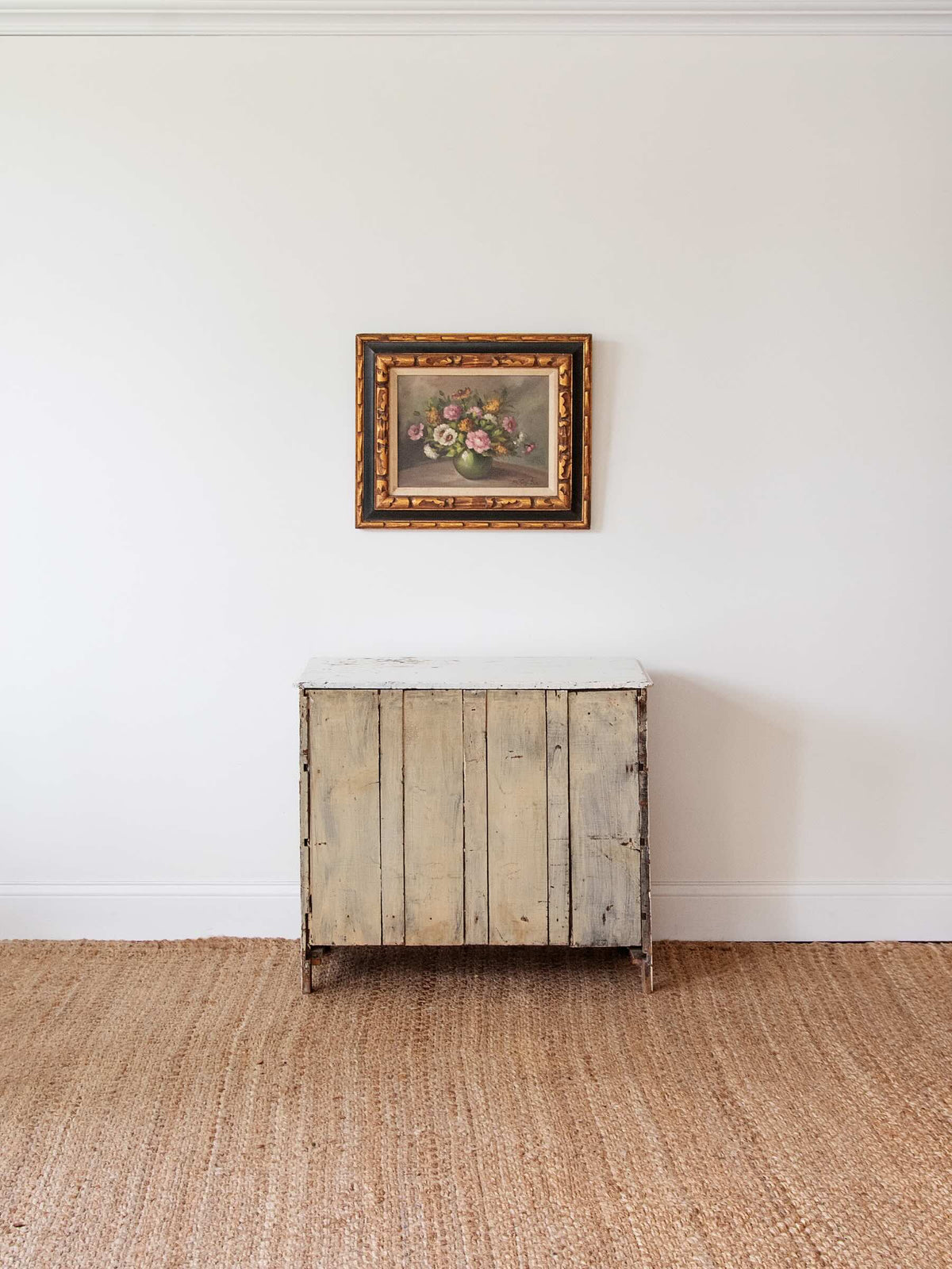 A rustic, weathered light beige wooden chest or cabinet with a white top, set against a white wall on a natural jute rug, with a framed floral painting hanging above it.