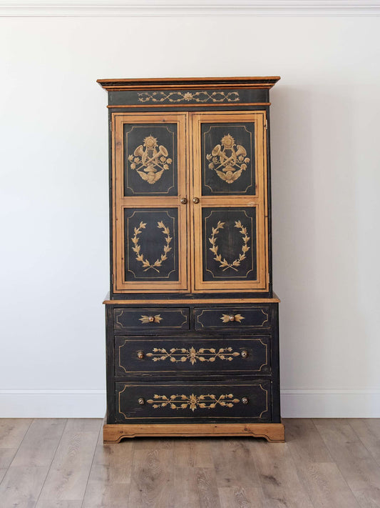 An antique black and gold cabinet with ornate decorative panels on the upper doors and drawer fronts, standing on a light wood floor against a white wall.
