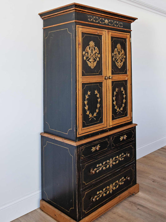 An antique black and gold cabinet with a two-door upper section and three drawers below, photographed in a room with white walls and light wood flooring.