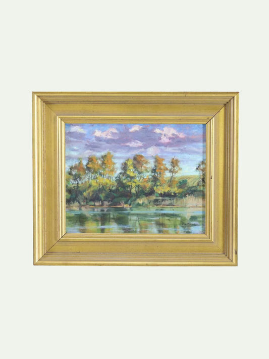 A framed landscape painting depicting trees by a lake under a cloudy sky, enclosed in a gold-colored frame against a light gray background.
