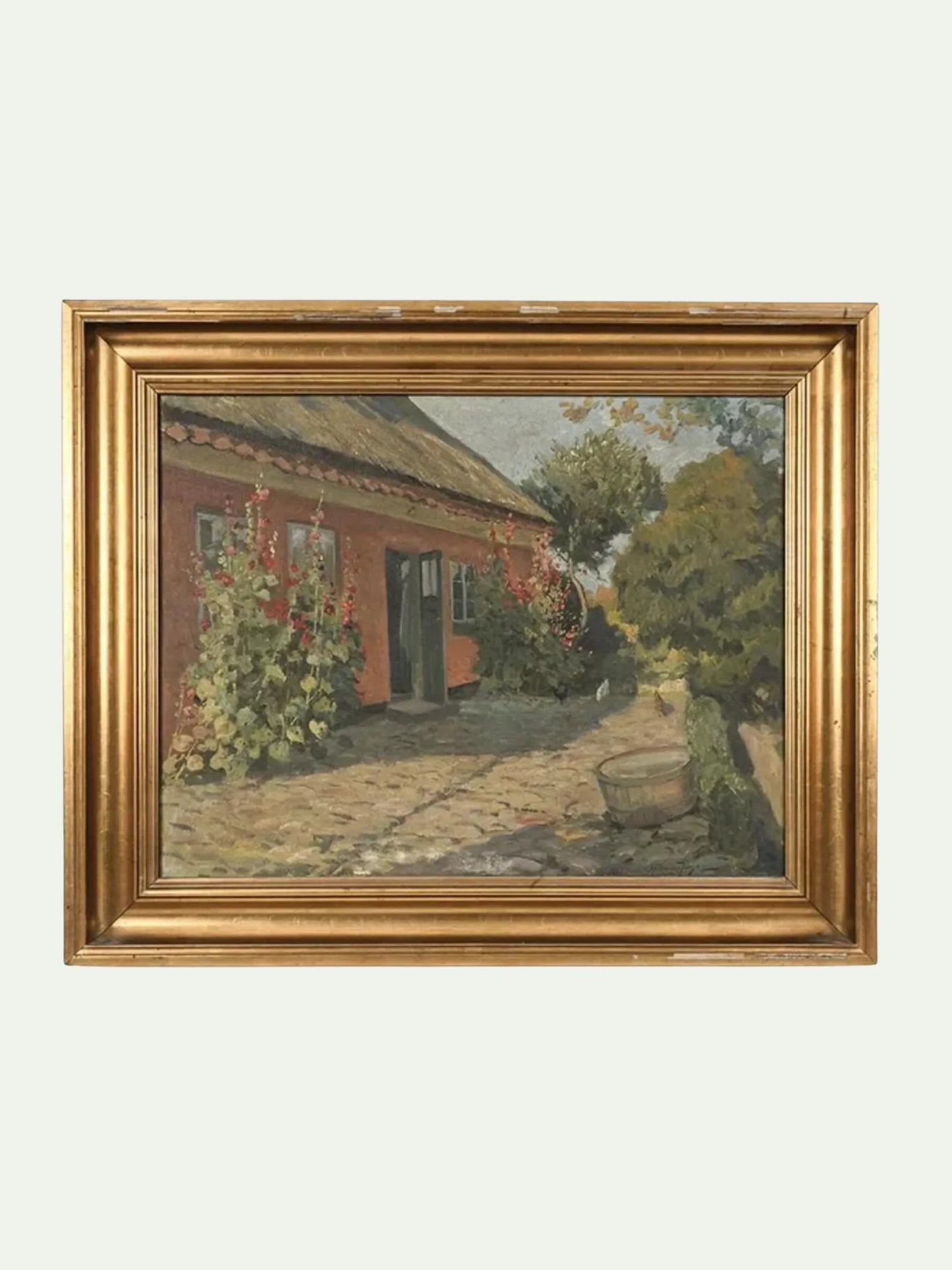 A framed landscape oil painting depicting a rustic red-orange cottage with a stone path, green trees, and climbing flowers, housed in an ornate gold frame against a light gray background.