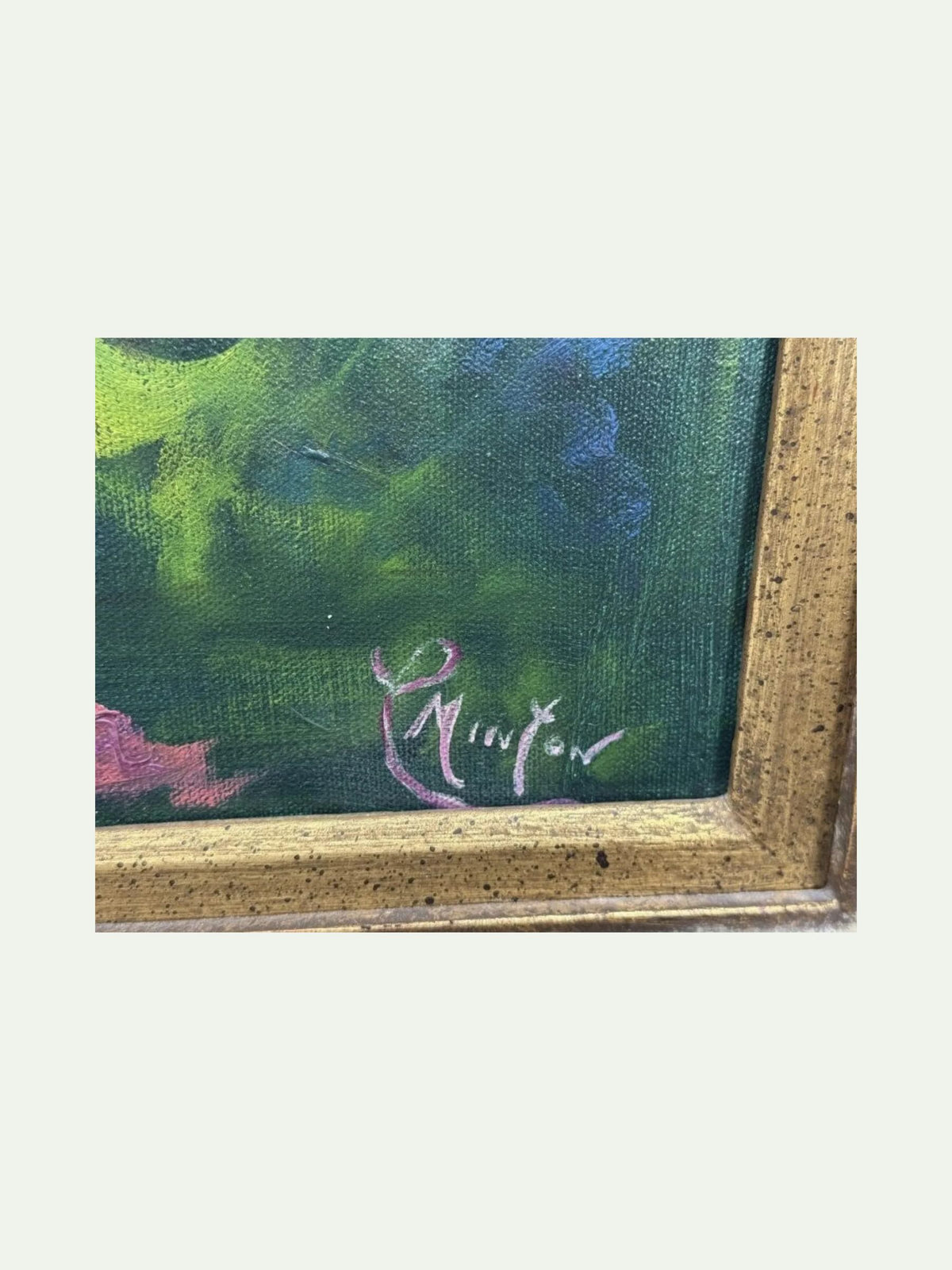 Close-up of a framed painting corner showing a textured green and blue painted surface with a pink signature and a speckled gold frame on a light background.