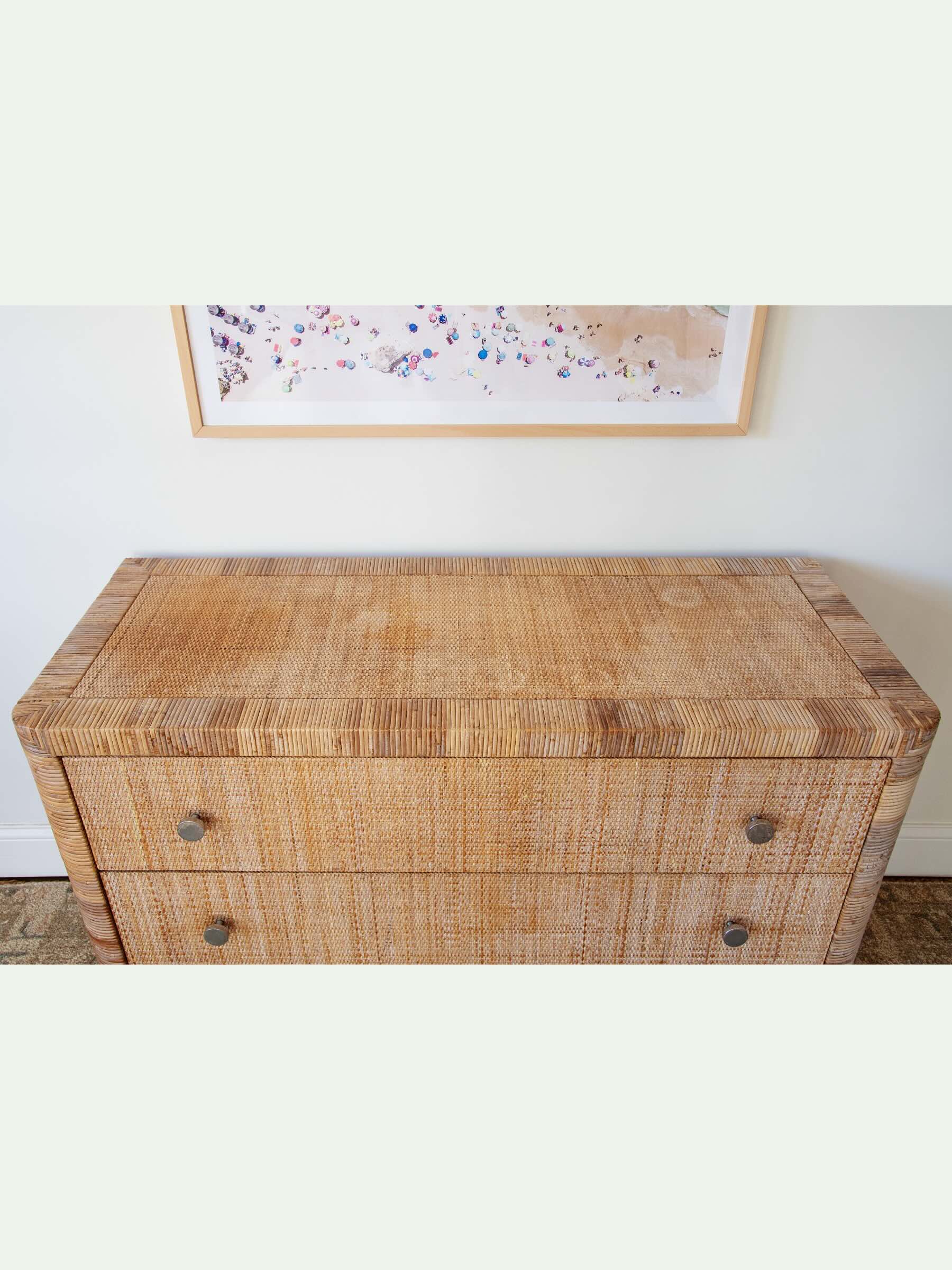 Top profile of wicker dresser with framed beach scene artwork on a white wall.