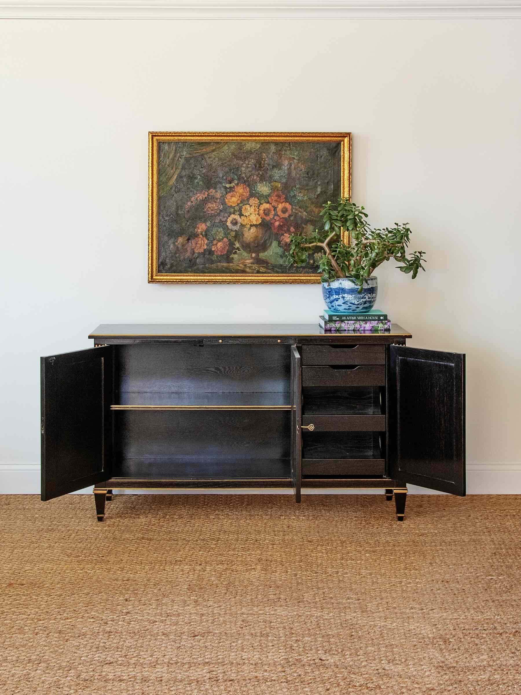 Black sideboard with open doors against a white wall with a framed painting and plant.