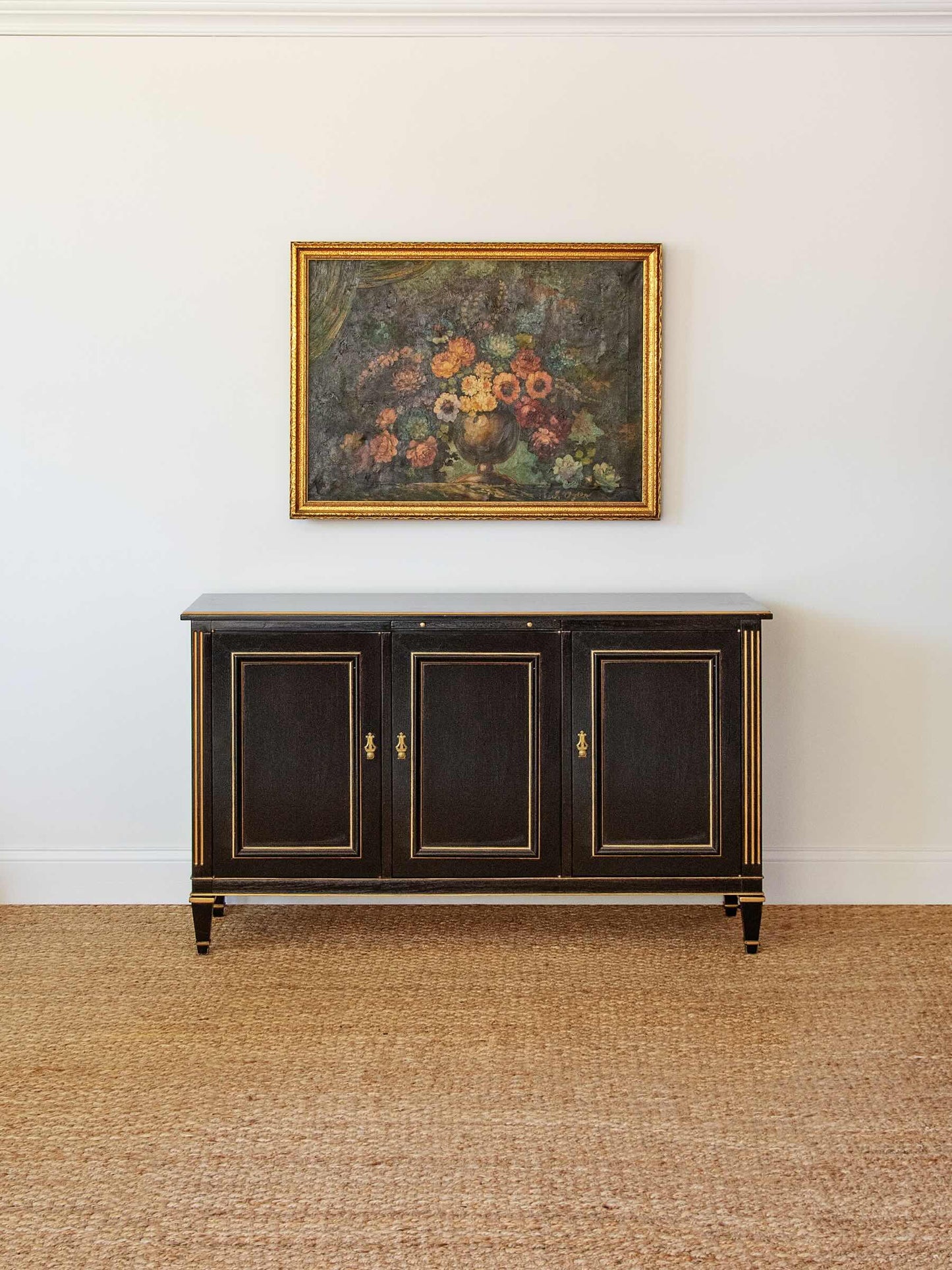 Dark wooden sideboard with gold accents against a white wall with a floral painting.