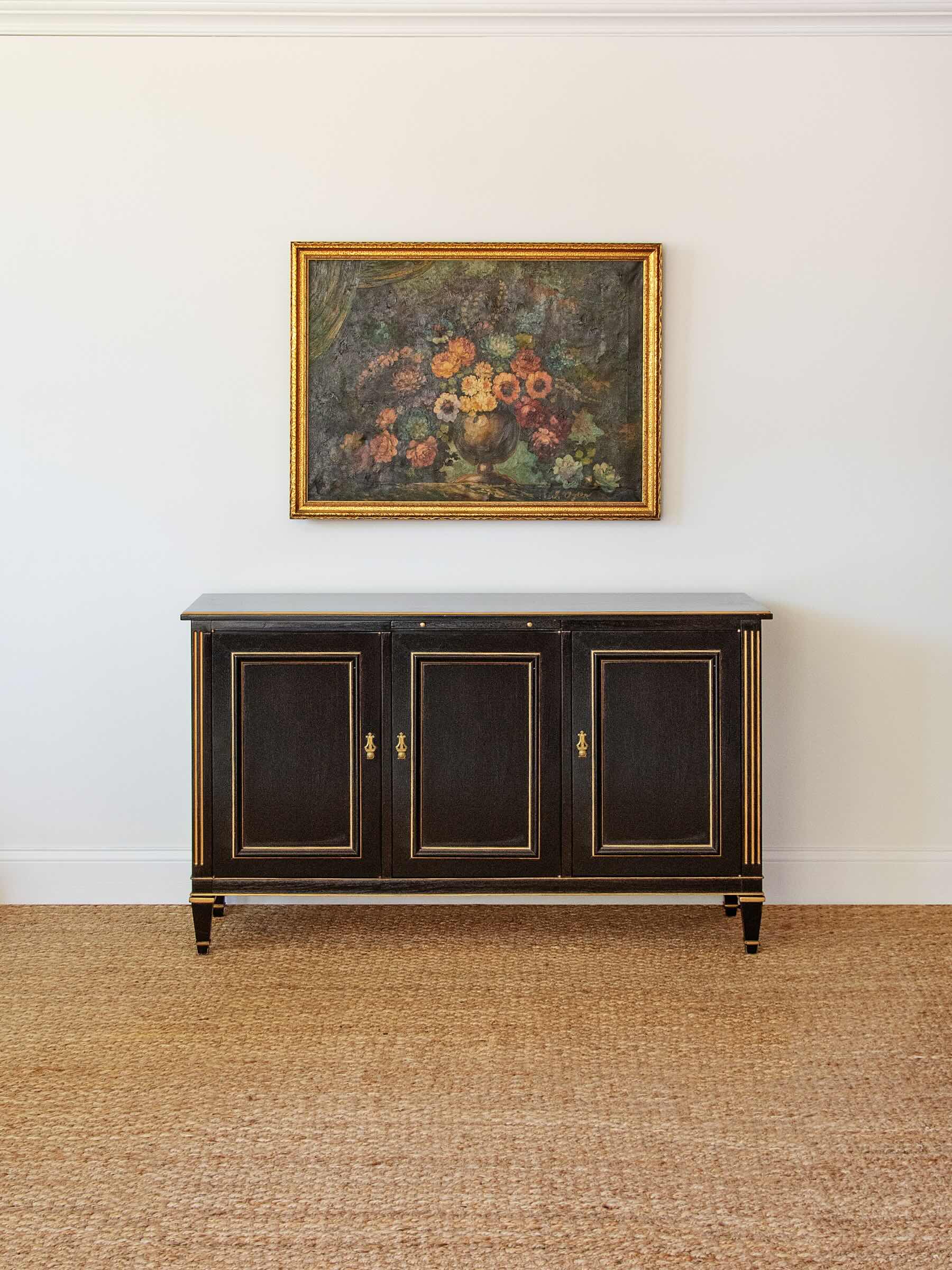 Dark wooden sideboard with gold accents against a white wall with a floral painting.