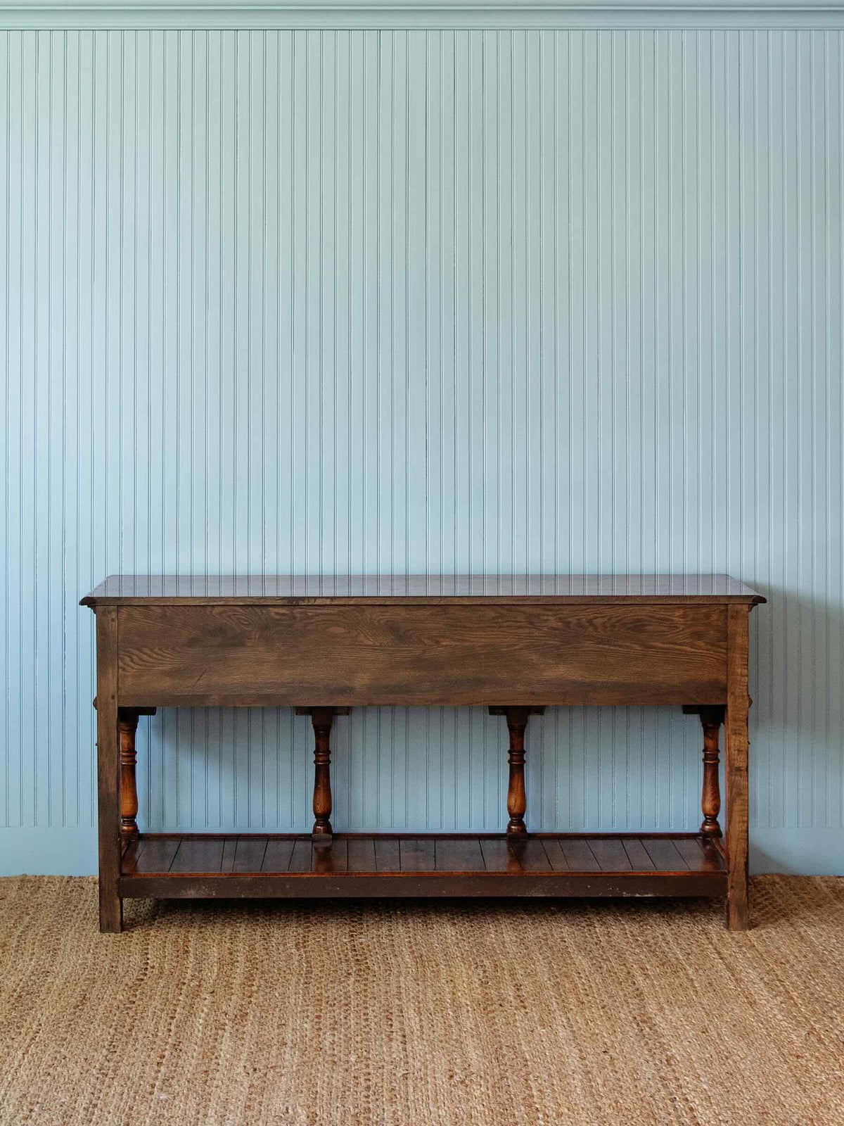A dark brown wooden console table with turned legs and a lower shelf, set against a light blue-beige beadboard wall and a natural jute rug.