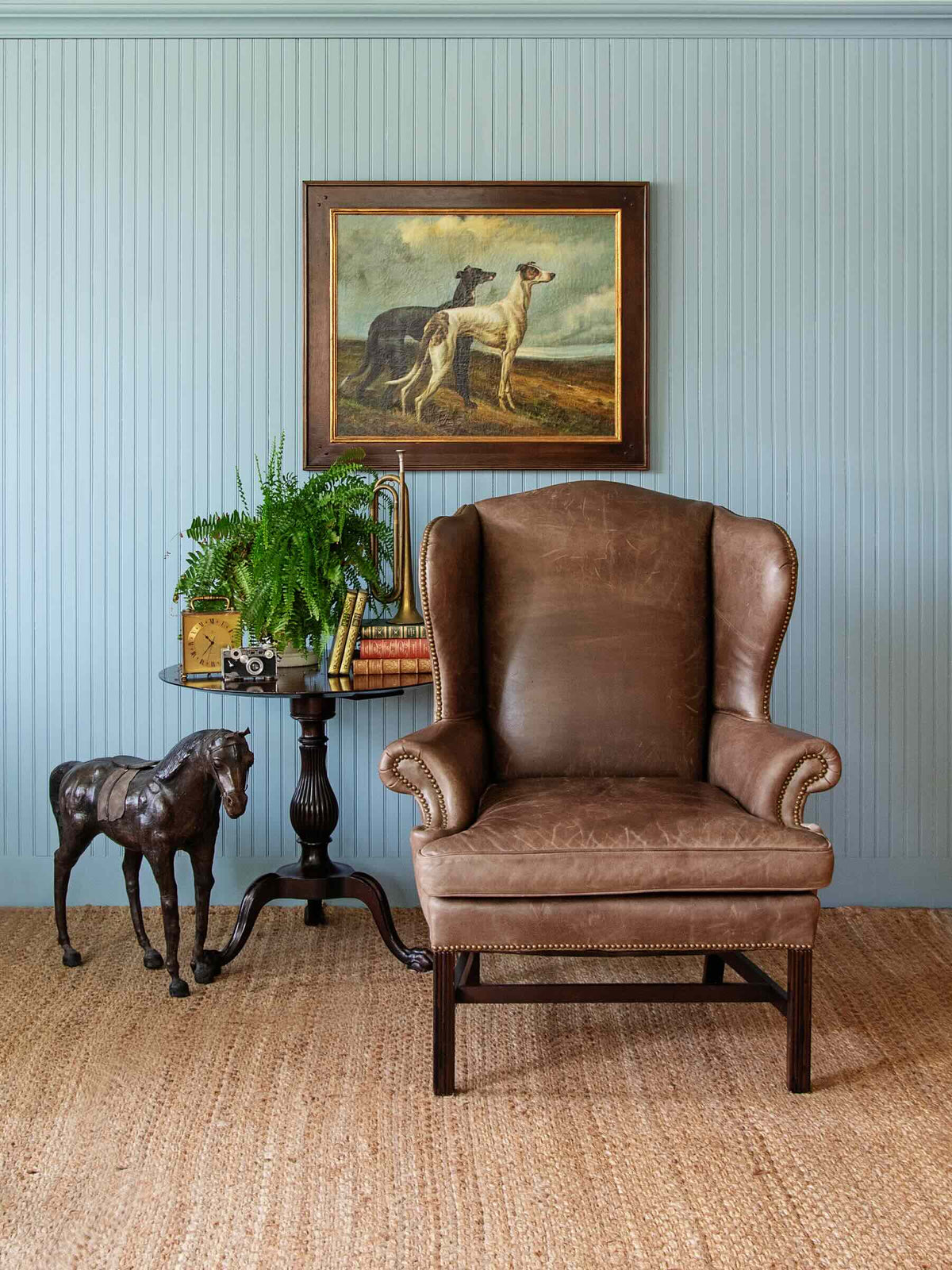 A brown leather wingback armchair in a styled room setting with a blue paneled wall, a framed painting of two dogs, a small side table with books and a plant, and a woven jute rug.