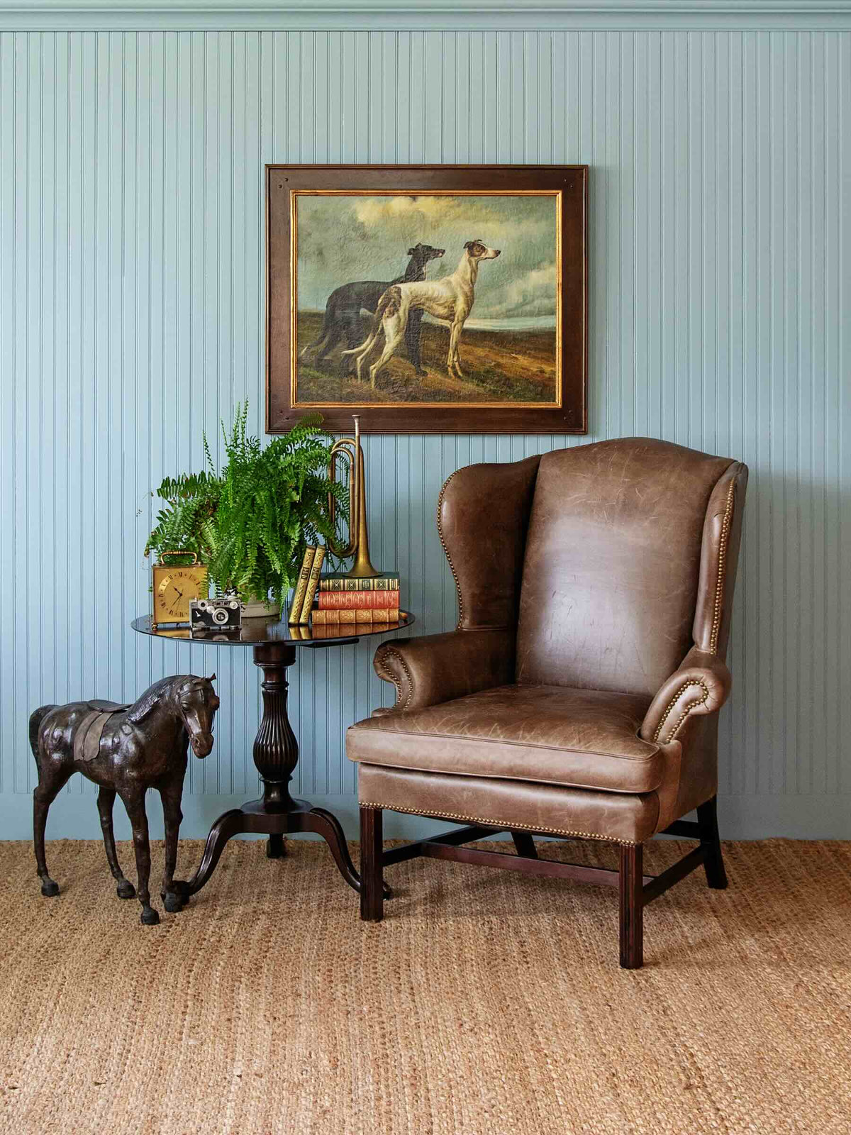 A brown leather wingback armchair in a styled room setting with a blue paneled wall, a framed painting of two dogs, a small side table with books and a plant, and a woven jute rug.