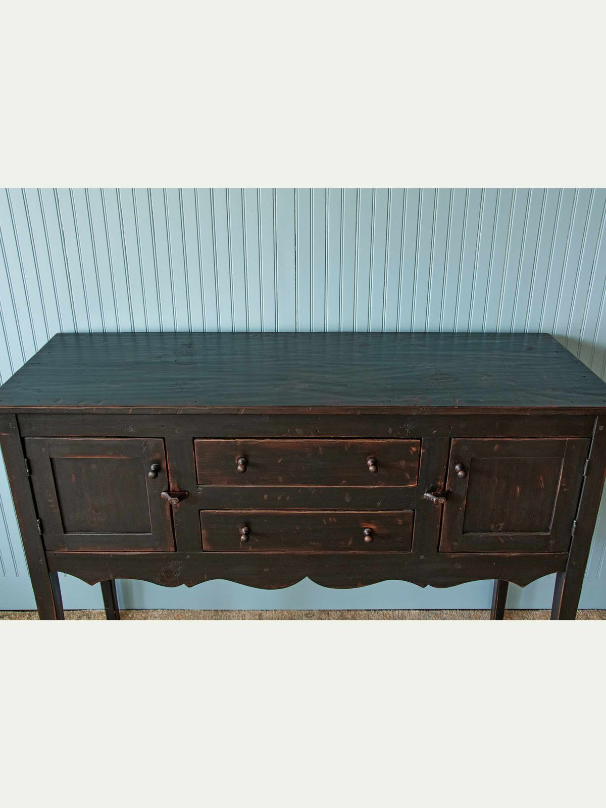 A dark brown wooden sideboard or buffet table with three drawers and cabinet doors, set against a light blue vertical wood-paneled wall.