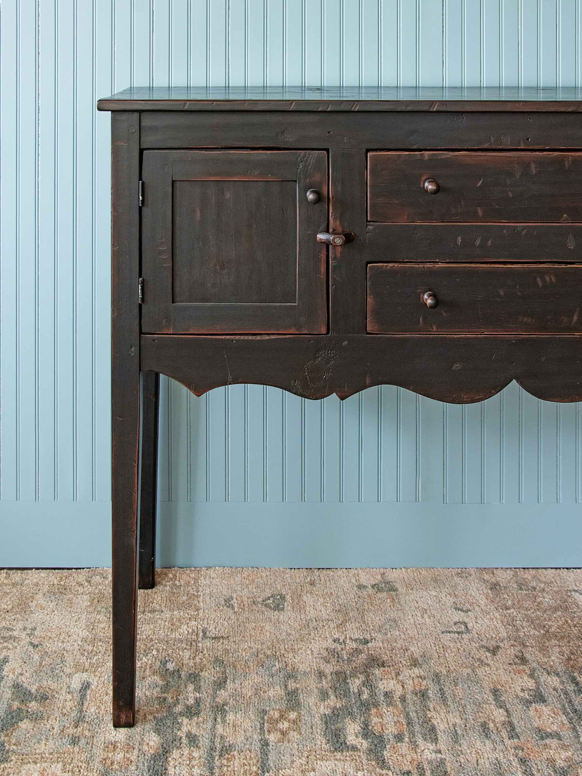 A dark stained wooden sideboard or buffet with a glass top, cabinet door, and drawers, set against a light blue paneled wall and a patterned rug.