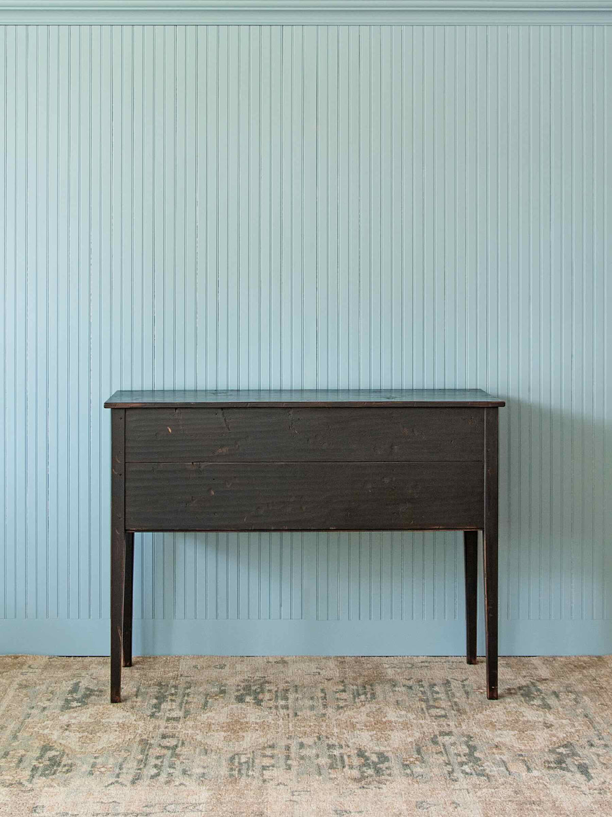A dark brown wooden console table with a glass top, set against a light blue vertical beadboard wall and a distressed beige rug.