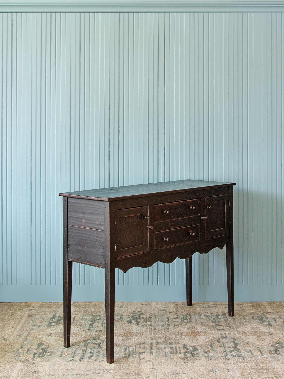 A dark brown wooden console table with three drawers and scalloped apron details, set against a light blue paneled wall and a distressed beige rug.