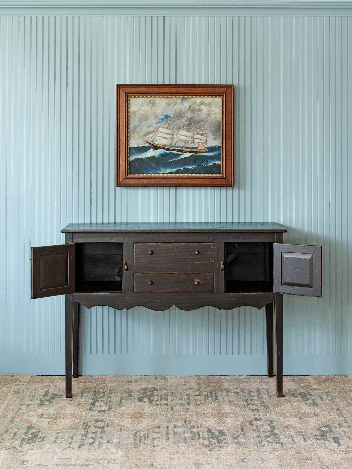 A dark wood console cabinet with open doors and three drawers, set against a light blue paneled wall with a framed painting above it.