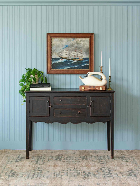A dark wood console table styled in a room setting against a light blue paneled wall, featuring a framed sailing ship painting, a white swan figurine, candles, books, and a green plant.