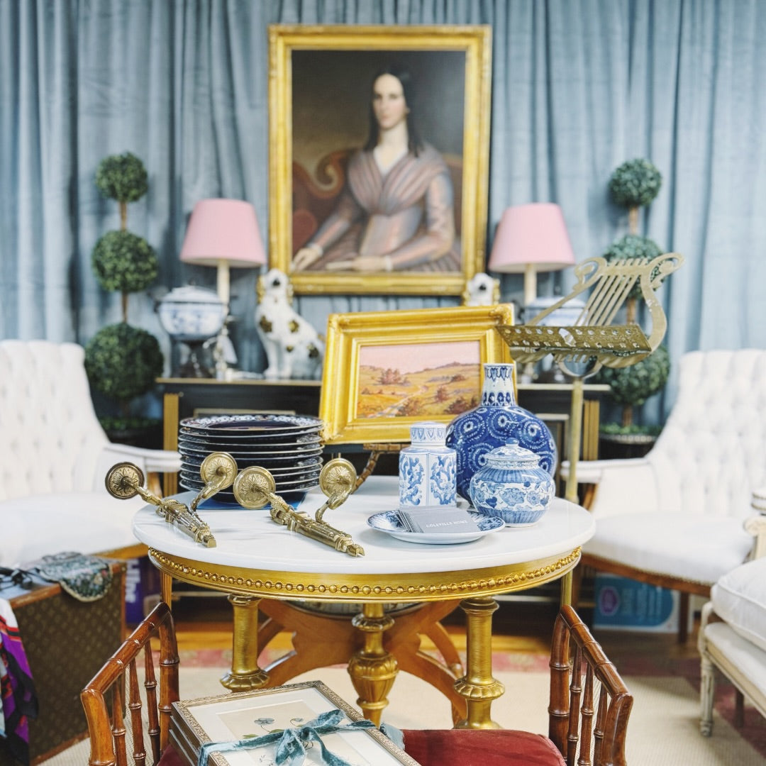 A styled interior scene with a round marble-top table in the foreground holding blue-and-white ceramic vases, plates, and brass-toned accessories, surrounded by white tufted armchairs, pink-lamp side tables, potted topiary trees, and a large framed portrait on blue curtains in the background.